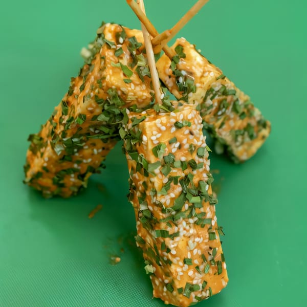 Three BBQ paneer skewers with sesame seeds and green herbs on a green background.