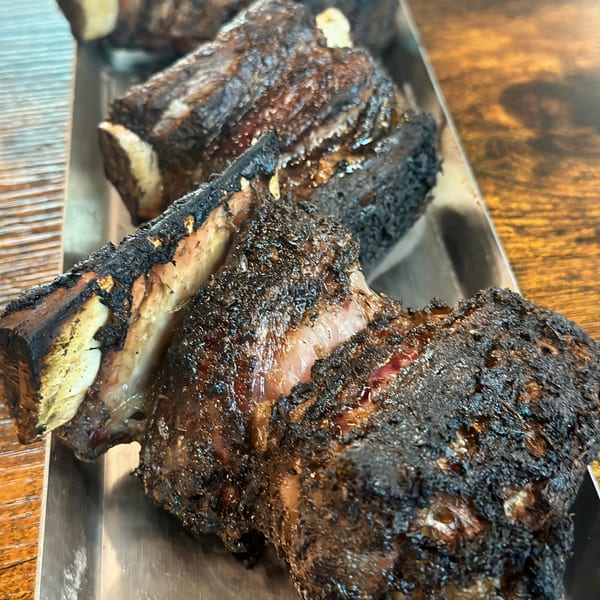 Close-up of several large, charred BBQ beef ribs on a metal tray, showcasing crispy bark and tender meat.