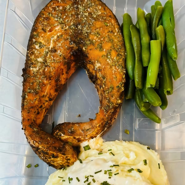 BBQ salmon steak with green beans and mashed potatoes, garnished with chives, in a takeout container.