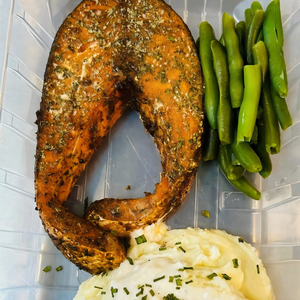 BBQ salmon steak with green beans and mashed potatoes, garnished with chives, in a takeout container.